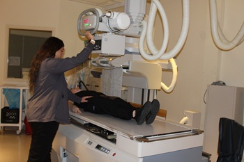 Picture of a female Nurse standing behind a female patient that is standing straight up, getting an X-RAY done.